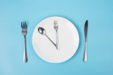 Mavi zemin üzerinde bıçak, kaşık ve çatal olan beyaz seramik plaka. Aralıklı oruç, Ketogenik diyet, kilo kaybı, yemek planı ve sağlıklı gıda konsepti