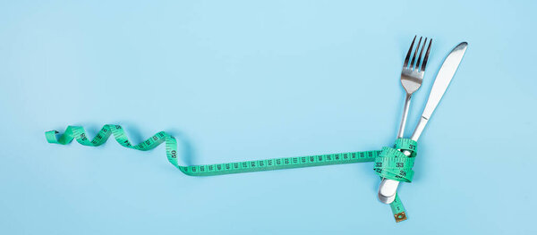Top view Green Measuring tape wrapped around fork and knife on blue background. dieting, weight loss, obesity and food control concept