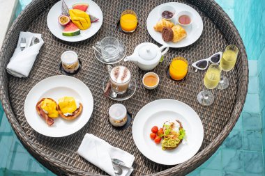 Lüks otel veya tropikal tatil köyünde yüzme havuzunda yüzen kahvaltı tepsisi, meyve; mango, karpuz ejderhası ve tutku meyvesi, yiyecek, ekmek, kahve, çay, şarap ve portakal suyu. Egzotik yaz