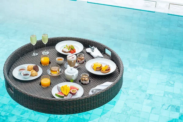 Floating Breakfast tray in swimming pool at luxury hotel or tropical resort villa, fruits; mango, watermelon dragon and passion fruit, food, bread, coffee, tea, wine and orange juice. Exotic summer