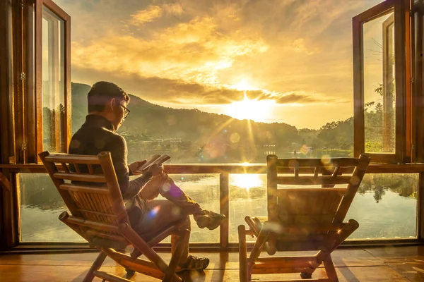 young man reading book near window and watching lake view at coffee shop in the morning sunrise, Ban Rak Thai village, Mae Hong Son province, Thailand. Travel concept