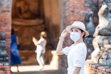 Ayutthaya 'daki Wat Chaiwatthanaram Tapınağı ziyareti sırasında cerrahi yüz maskesi takan, COVID-19 salgınını koruyan beyaz elbiseli turist kadın. Yeni normal, güvenli seyahat ve Tayland seyahat kavramı