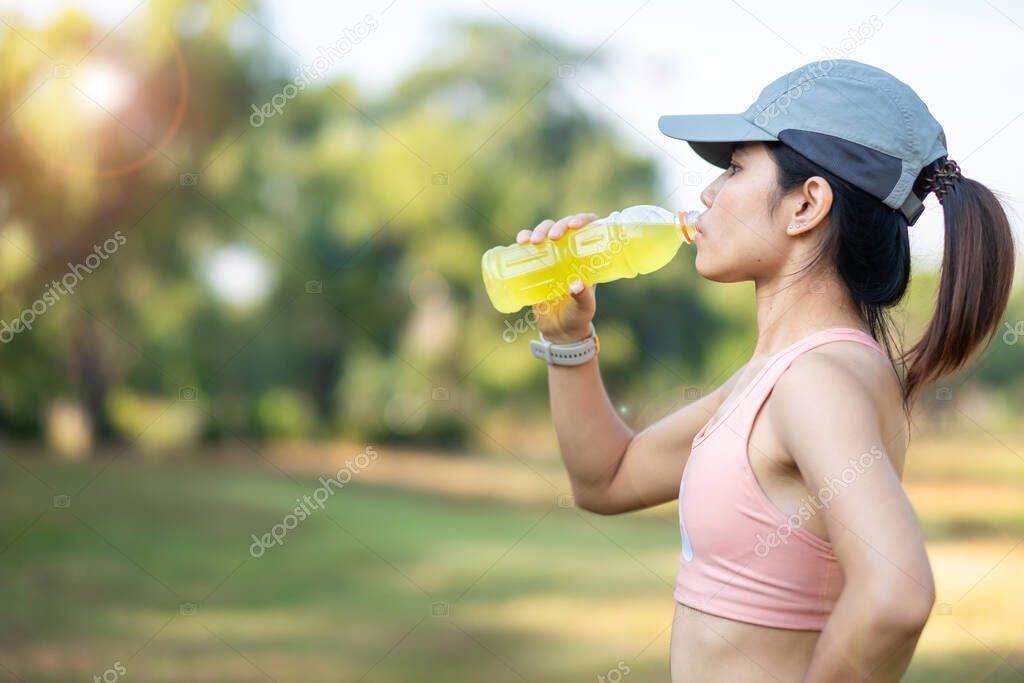 young fitness woman drinking energy water during stretching muscle in the park outdoor, asian athlete running and exercise in morning. Sport and wellness concept