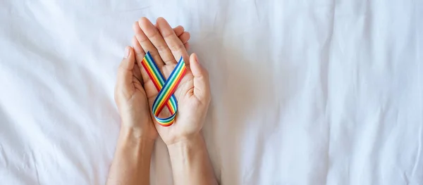 hand showing LGBTQ Rainbow ribbon on white bed background. Support Lesbian, Gay, Bisexual, Transgender, Queer community and Rights concept