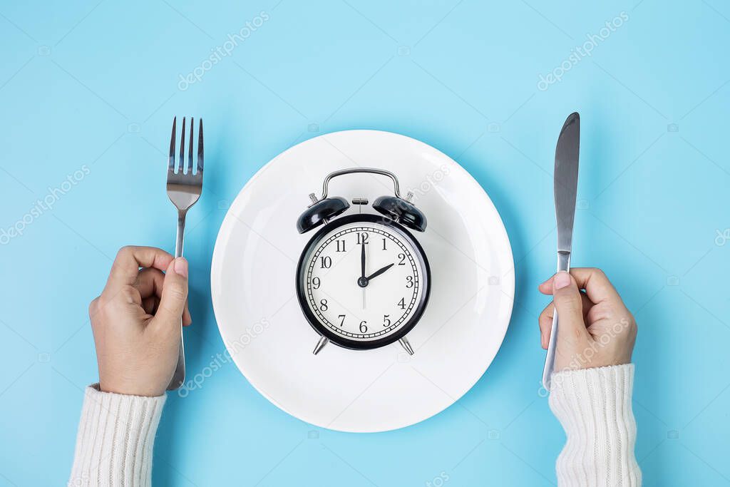 Hands holding knife and fork above alarm clock on white plate on blue background. Intermittent fasting, Ketogenic dieting, weight loss, meal plan and healthy food concept