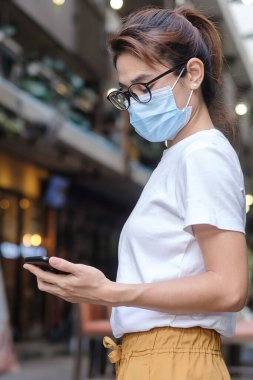 Tıbbi yüz maskesi takan ve gece pazarında yürürken akıllı telefon kullanan bir kadın, Coronavirus veya Corona Virüs Hastalığını (Covid-19) engeller. Sağlık, yaşam konsepti