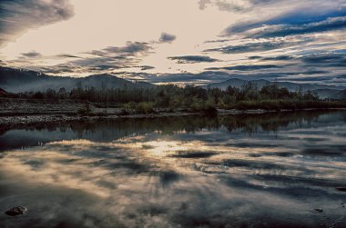 Aydınlatılmış tepeleri olan yüksek dağları olan güzel bir manzara, dağ gölündeki taşlar, yansıma, mavi gökyüzü ve gündoğumunda sarı güneş ışığı.