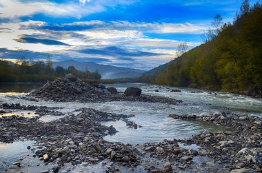 Aydınlatılmış tepeleri olan yüksek dağları olan güzel bir manzara, dağ gölündeki taşlar, yansıma, mavi gökyüzü ve gündoğumunda sarı güneş ışığı.