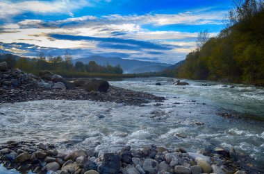 Aydınlatılmış tepeleri olan yüksek dağları olan güzel bir manzara, dağ gölündeki taşlar, yansıma, mavi gökyüzü ve gündoğumunda sarı güneş ışığı.