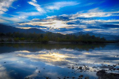 Aydınlatılmış tepeleri olan yüksek dağları olan güzel bir manzara, dağ gölündeki taşlar, yansıma, mavi gökyüzü ve gündoğumunda sarı güneş ışığı.