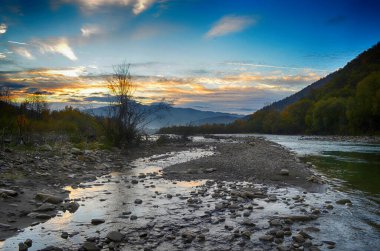 Aydınlatılmış tepeleri olan yüksek dağları olan güzel bir manzara, dağ gölündeki taşlar, yansıma, mavi gökyüzü ve gündoğumunda sarı güneş ışığı.