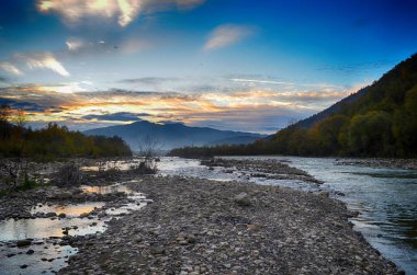 Aydınlatılmış tepeleri olan yüksek dağları olan güzel bir manzara, dağ gölündeki taşlar, yansıma, mavi gökyüzü ve gündoğumunda sarı güneş ışığı.