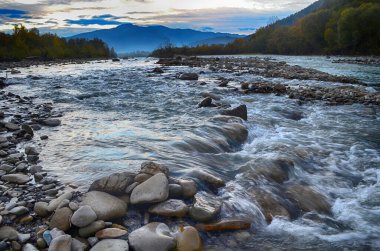 Aydınlatılmış tepeleri olan yüksek dağları olan güzel bir manzara, dağ gölündeki taşlar, yansıma, mavi gökyüzü ve gündoğumunda sarı güneş ışığı.