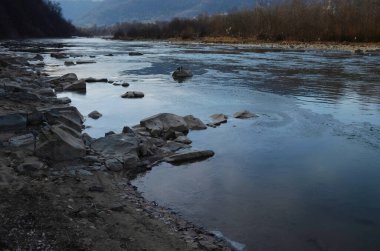Kış manzarasında donmuş bir nehir. Ormanlı kış manzarası, bulutlu gökyüzü ve güneş.