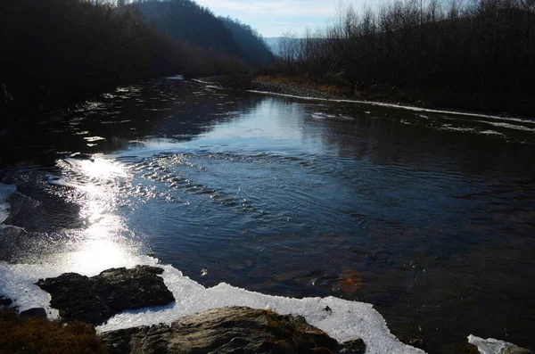 A frozen river in a wintry landscape. Winter landscape with forest, cloudy sky and sun.