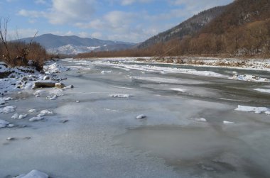 Kış manzarasında donmuş bir nehir. Ormanlı kış manzarası, bulutlu gökyüzü ve güneş.