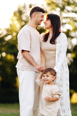 Sevgi dolu genç ebeveynler gülümseyen bebekleri dışarıda sıcak bir altın saatin tadını çıkarırken kucaklaşırlar. Doğal aile anları bir park ortamında sevgi, birliktelik, ebeveynlik ve neşeli bir çocukluk geçirir..