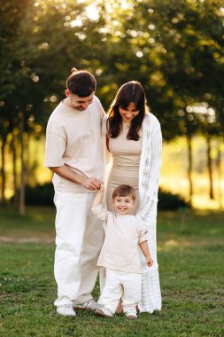 Mutlu, genç bir aile, altın saatinde güneşli bir parkta yürüyor. Aileler gülümseyen bebeklerinin elini tutuyor. İçtenliklerini ve yakınlıklarını ifade ediyorlar..