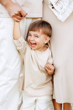 Neşeli bir çocuk anne ve babasının elini tutarak gülerken anne ve babasının arasında nötr bir tonda duruyor. Sıcak, samimi bir aile anı. Aşkı, güvenliği, masumiyeti ve neşeli bağlantıyı aktaran..