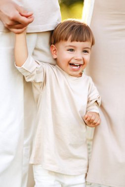 Gülümseyen çocuk sıcak bir açık havada aile portresinde ebeveynlerin ellerini tutuyor, tarafsız kıyafetlerle neşeyle gülüyor ve sevgi, güven, neşeli çocukluk anları ve yakın aile bağlarını paylaşıyor..