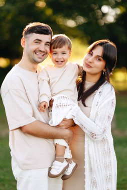 Gülümseyen ebeveynler neşeli bebeklerini sıcak bir açık havada altın saat portresinde tutarlar. Sevgiyi, birlikteliği ve güneşli bir parkta rahat aile anlarını aktarırlar..