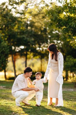 Genç ebeveynler gülümseyen çocuklarına, güneşli bir parkta yürüdükleri altın saat boyunca, sıcak aile bağları, nazik ebeveynlik, neşe, şefkat ve rahat açık hava yaşam tarzı anlarını yakalamalarında yardımcı olurlar..