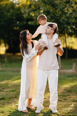 Genç ve mutlu bir aile, bir parkta altın saat güneş ışığının tadını çıkarırken, baba, oğlunu omuzlarında taşır ve anne sevgi dolu, sevgi dolu, birliktelik ve ebeveynlik bağıyla ona bakar..