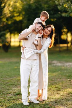 Genç ebeveynler kucaklaşıp gülümserken, bebekleri güneşli bir parkta babalarının omuzlarına biniyor, sıcak aile sevgisini yakalıyor, neşeli ebeveynlik anları yakalıyor.