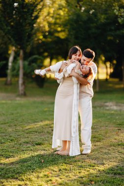Genç ebeveynler, ağaçlar ve yeşil çimlerle çevrili altın saat boyunca kızlarıyla birlikte neşeli bir şekilde güneşli bir parkta gülüşlerini ve sıcak aile anlarını paylaşıyorlar..