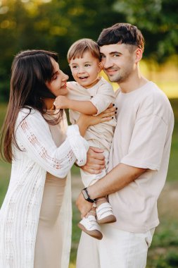 Genç ebeveynler, güneşli bir parkta neşeli bebeklerini kucaklarken ve kucaklarken gülümserler. Sıcak, sevgi, birliktelik ve aile bağlarını dışardaki rahat altın saat boyunca ifade ederler..