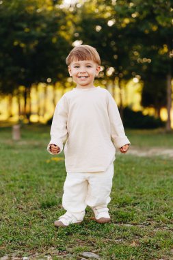 Güler yüzlü çocuk, altın saatinde güneşli bir parkta çimlerin üzerinde, açık renkli giysiler ve spor ayakkabılar giyerek duruyor. Neşeli, neşeli ve masum ruh hali. Sıcak doğal arka plan ve bokeh ağaçları..