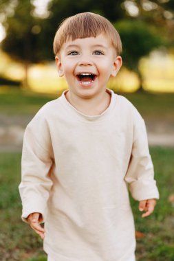 Güneşli bir parkta, açık havada çekilmiş bir resim çekilirken gülüp gülümseyen, açık tişörtlü, neşeli, kaygısız çocukluk anılarının tadını çıkaran mutlu bir çocuk..