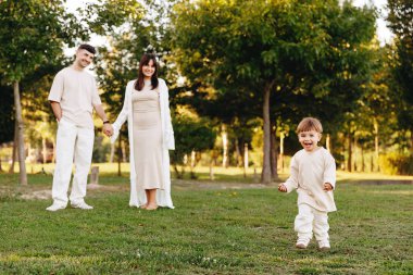 Mutlu, genç bir aile parkta güneşli bir günün tadını çıkarırken neşeli bir çocuk çimenlerin üzerinde koşuyor ebeveynler el ele tutuşup gülümsüyor, neşeli bağları ve kaygısız açık hava anlarını yakalıyor..