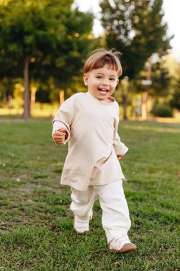 Mutlu bir çocuk altın saatinde çimenli bir parkta koşar ve hafif günlük kıyafetlerle güler. Doğal açık hava portresi neşeli hareketleri, çocukluk enerjisini ve neşeli yaz atmosferini yakalar..