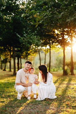 Üç anne, baba ve çocuktan oluşan gülümseyen genç bir aile, açık havada sevgi, neşe ve beraberlik taşıyan yapraklı bir parkta altın bir günbatımında sıcak bir kucaklaşmayı paylaşıyor..