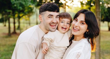 Genç ebeveynler güneşli bir parkta birlikte gülerken neşeli çocuklarını kucaklarlar, sıcaklığı, birlikteliği, aile bağlarını ve kaygısız açık havada doğal yeşil bir ortamda oynarlar..