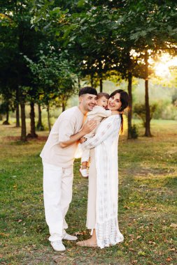 Genç ebeveynler gülümseyen bebeklerini güneşli bir parkta kucaklıyor, altın saatinde ağaçların arasında gezinirken sıcak, sevgi ve beraberlik dolu bir anı paylaşıyorlar..