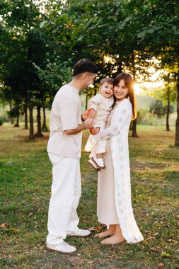 Üç anne, baba ve çocuktan oluşan gülümseyen genç bir aile, açık havada sevgi, neşe ve beraberlik taşıyan yapraklı bir parkta altın bir günbatımında sıcak bir kucaklaşmayı paylaşıyor..