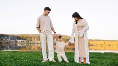 Sevgi dolu ebeveynler, altın saatinde sakin bir göl kıyısında yürür. El ele tutuşur ve doğada huzurlu bir anın tadını çıkarırlar. Sıradan bir giysi ve sıcak bir güneş ışığı huzurlu bir aile portresi oluşturur..