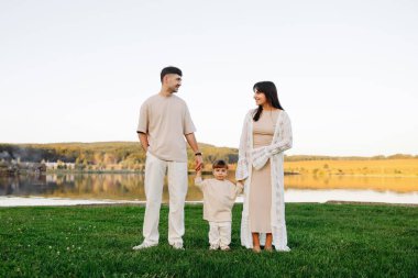 Genç ebeveynler gün batımında sakin bir gölün kenarında yeşil çimlerin üzerinde durmuş, bebeklerinin ellerini tutuyor ve gülümsüyorlar. Doğal ışık, rahat giysiler ve sıcak tonlar sevgiyi, birlikteliği ve huzurlu aile yaşam tarzını ifade eder..