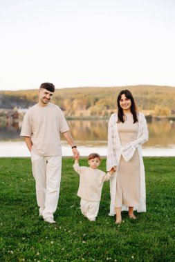 Genç ebeveynler, sakin bir gölün kenarındaki yeşil çimlerde yürüyorlar, eşgüdümlü nötr giysiler giyiyorlar, gülümsüyorlar, aile birliğini, sıcaklığı, ebeveynliği ve açık hava yaşam tarzını aktarıyorlar..