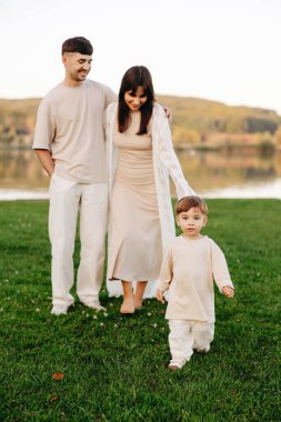 Genç ebeveynler, sakin bir gölün kenarındaki yeşil çimlerde yürür ve açık havada huzurlu bir anı paylaşırlar. Tarafsız tonlar ve yumuşak akşam ışığı huzurlu bir aile portresi oluşturur..