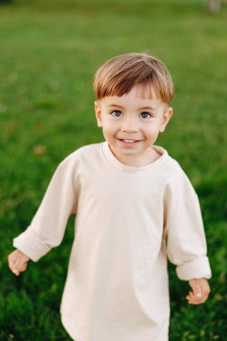 Gülümseyen küçük bir çocuk yeşil çimlerin üzerinde krem rengi bir gömlekle kameraya bakıyor. Doğal açık hava portresi neşeli masumiyeti, neşeli enerjiyi, sıcak ışığı ve günlük aile çocukluğu anlarını yakalıyor..