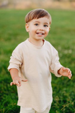 Yeşil çimlerin üzerinde duran, bej renkli uzun kollu gömlek giyen ve kameraya bakan gülümseyen çocuk. Sıcak, neşeli açık hava portresi neşe, masumiyet, çocukluk ve aile anlarını aktarıyor..