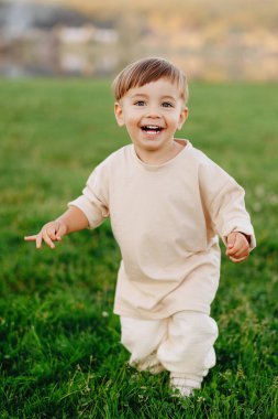 Mutlu çocuk, güneşli çimenlik bir arazide, tarafsız bej giysilerle koşuyor, gülüyor ve öne doğru uzanıyor. Doğal açık hava portresi neşeyi, oyunculuğu, masumiyeti ve tasasız çocukluk keşiflerini yakalar..