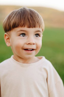 Kase kesimi olan gülümseyen çocuk, güneş ışığında yeşil bir alanda, masumiyet, merak ve neşe saçan bir çocuğa bakıyor. Yumuşak doğal ışık ve sığ alan derinliği samimi ve samimi bir portre oluşturur..