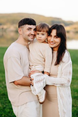 Genç ebeveynler, küçük oğullarını göl kenarındaki bir parkta tutarken gülümsüyor. Sıcak, samimi bir aile anını paylaşıyorlar. Altın saat boyunca, doğal bir bağ ve sevgiyle..