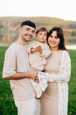 Gülümseyen genç aile göl kenarındaki bir çayırda yan yana durur, anne babalar yeni yürümeye başlayan çocuklarını yumuşak, nötr giysiler içinde tutarlar, sıcaklığı, birlikteliği, ebeveynliği ve açık hava yaşam tarzını altın saat aydınlığında aktarırlar..