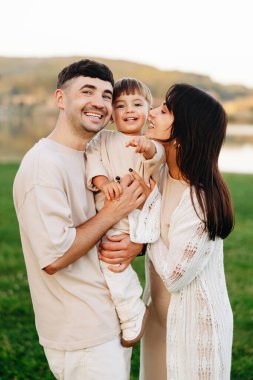 Mutlu genç ebeveynler göl kenarında gülümseyen bebekleriyle sarılıp oynuyorlar. Sevgi dolu anları, kahkahaları ve sıcak aile bağlarını doğal ve sıradan bir yaşam tarzında paylaşıyorlar..