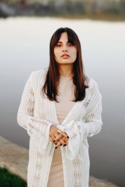 Beyaz örülü hırka ve bej bir elbise giyen genç bir kadın, alacakaranlıkta sakin bir gölün kenarında poz veriyor, sakin, derin düşüncelere dalmış bir şekilde dış mekan tarzı bir portrede doğal güzelliği yansıtıyor..